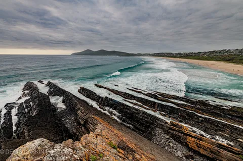 Layered rust-streaked shale and orange lichen in the foreground lead toward breaking waves and a sandy beach arc under a textured sky.