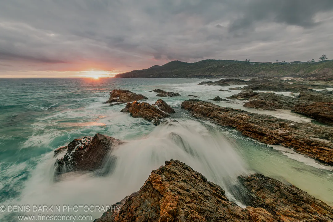 Wet sandstone formations on the Forster coast at dawn, with long-exposure surf and a hilly headland in the mist.