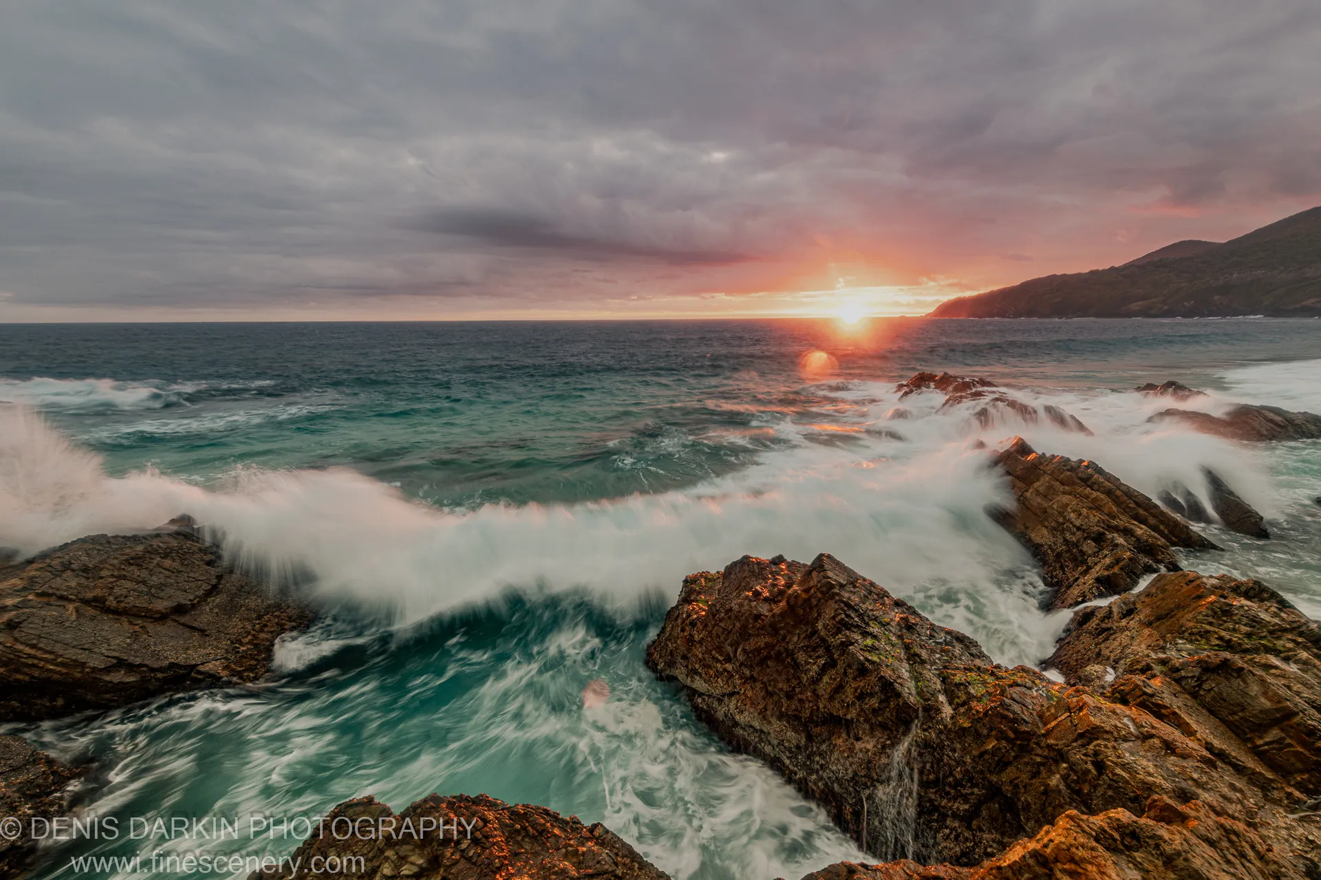 A breaking wave moves around rocky outcrops on the Forster coast at dawn, with orange light along the horizon below grey clouds.