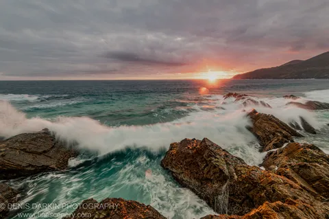 A breaking wave moves around rocky outcrops on the Forster coast at dawn, with orange light along the horizon below grey clouds.