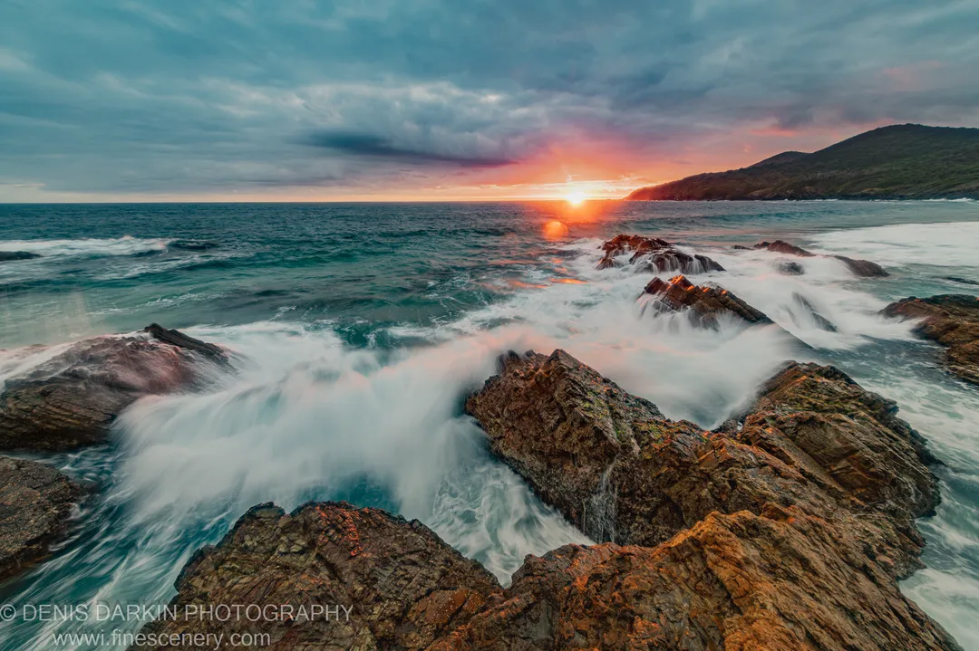 White water flows around dark angular boulders on the Forster coast at dawn, with storm clouds and warm light on the horizon.