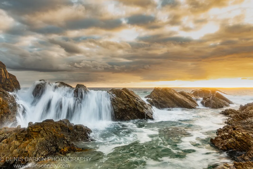 Layered sandstone outcrops on the Forster coast at dawn, with long-exposure waves and a sky shifting from grey cloud to warm light.