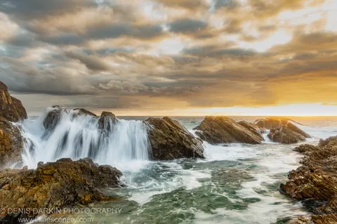 Layered sandstone outcrops on the Forster coast at dawn, with long-exposure waves and a sky shifting from grey cloud to warm light.