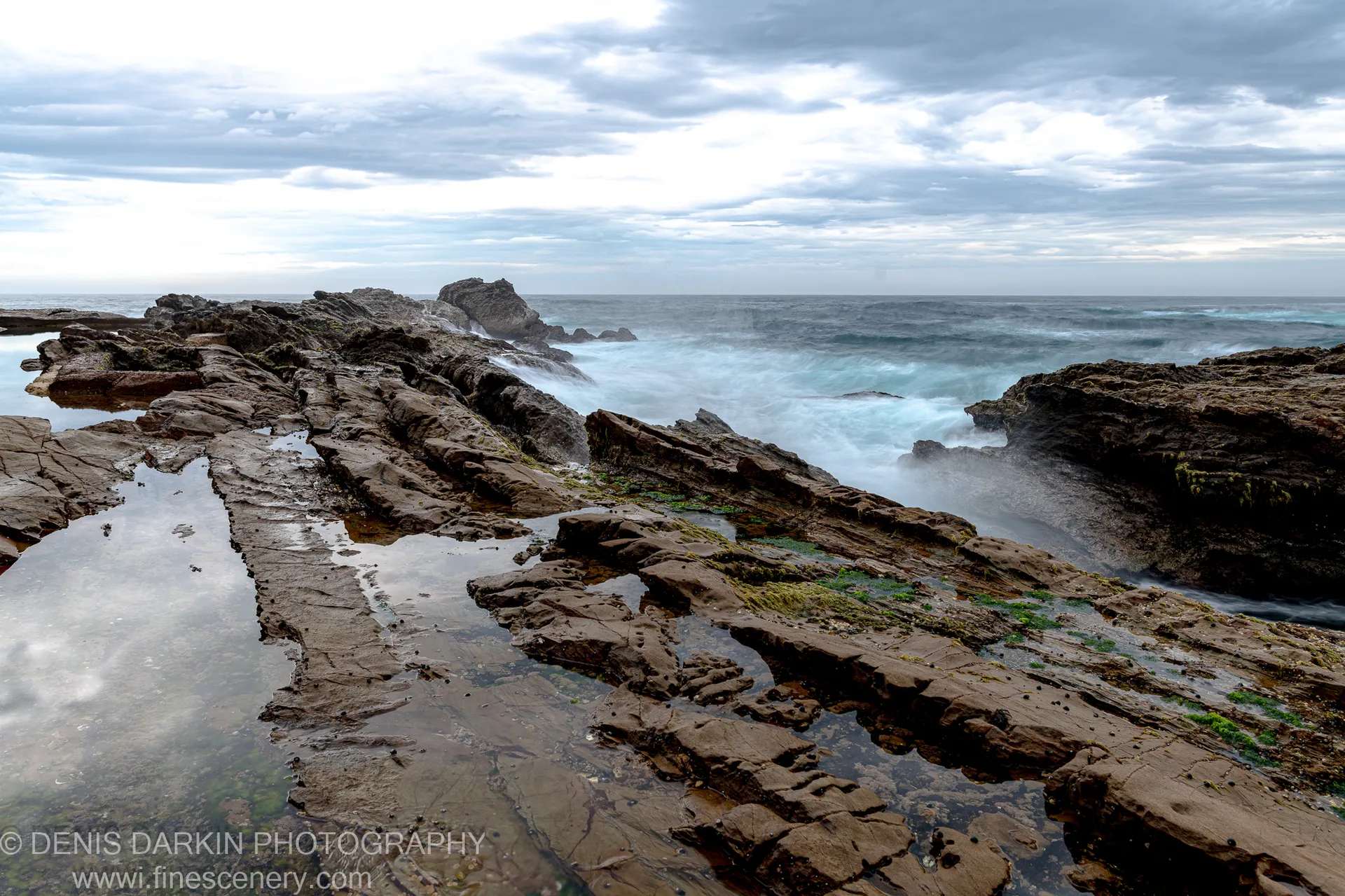 Wet layered rock platform with tide pools at dawn near Bermagui, grey clouds reflected in still water and surf breaking in the distance.