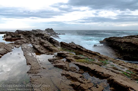 Wet layered rock platform with tide pools at dawn near Bermagui, grey clouds reflected in still water and surf breaking in the distance.
