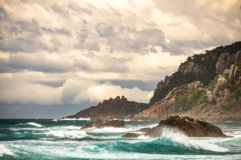 Rocky coastal headland on the New South Wales south coast, waves crashing against dark rocks below steep vegetated cliffs under a heavy, partially broken sky.