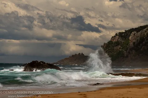 Powerful wave sending a large spray plume above dark cliff rocks at Bermagui, New South Wales, with heavy textured clouds filling the upper sky.