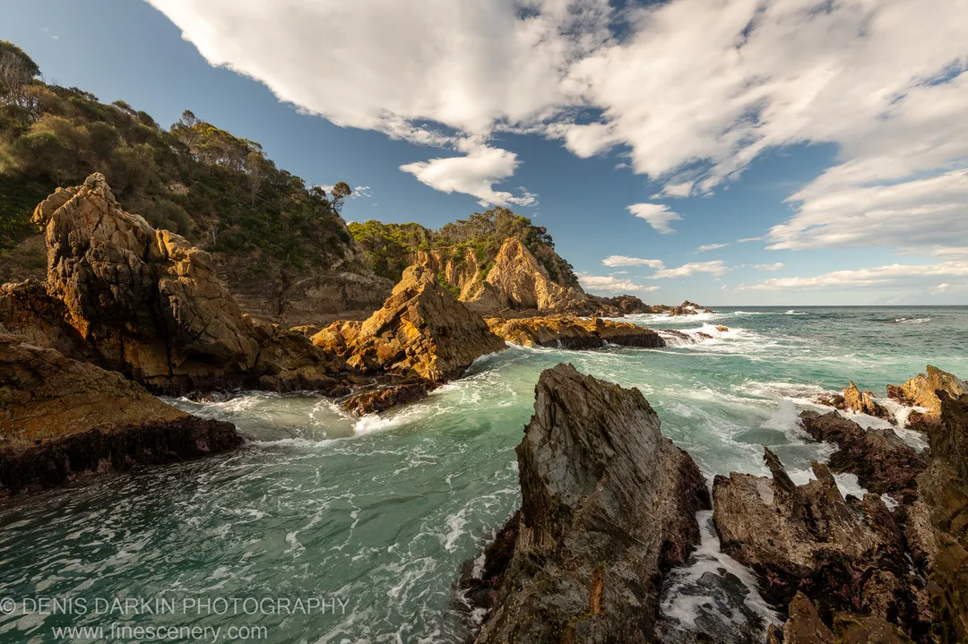 Golden-hour light on weathered sandstone cliffs at Bermagui where a freshwater estuary meets the ocean, scrub-covered hills rising against a blue sky with scattered clouds.