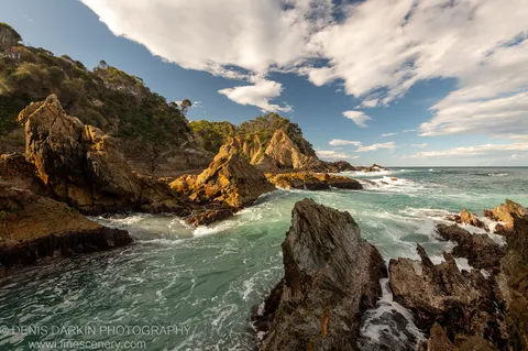 Golden-hour light on weathered sandstone cliffs at Bermagui where a freshwater estuary meets the ocean, scrub-covered hills rising against a blue sky with scattered clouds.