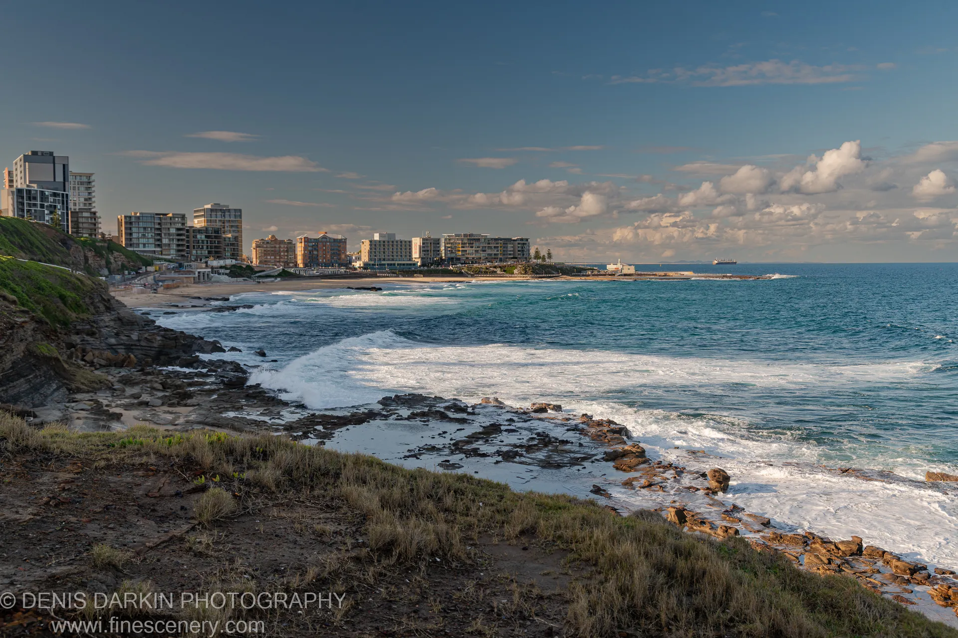 Layered coastal rock formations in the foreground contrast with Newcastle's high-rise buildings along the distant shoreline under a scattered cumulus sky.