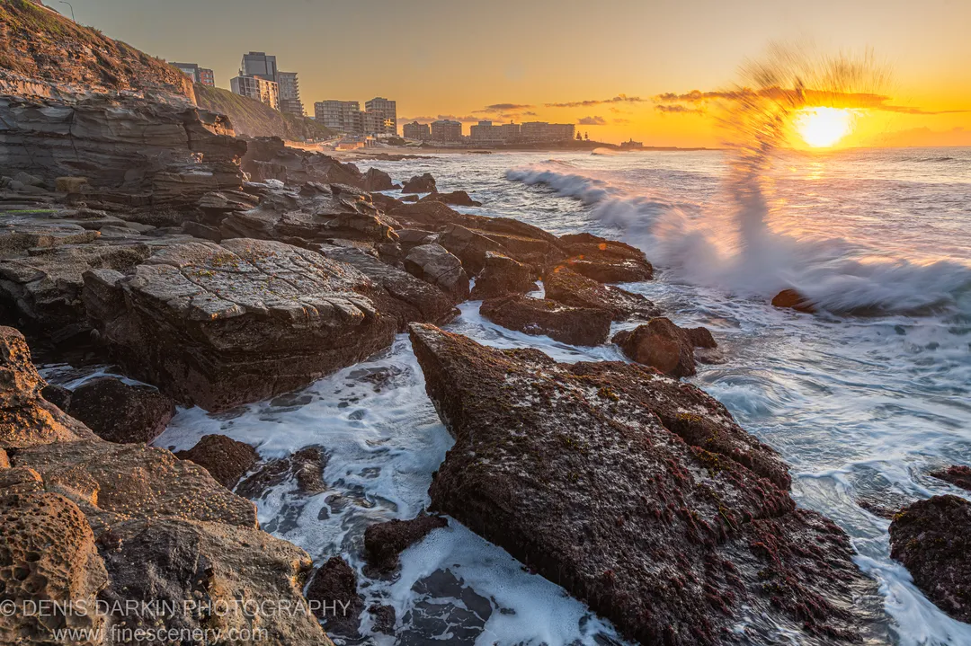 Moss-covered sandstone formations catch warm dawn light at Newcastle's shoreline, with ocean foam at their base and the city skyline visible on the distant cliffs.