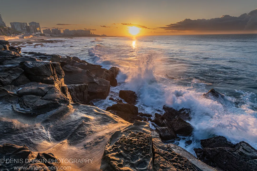 Honeycomb-weathered limestone at the Newcastle shoreline at dawn, a wave breaking at its base and the sun's reflection across the water.