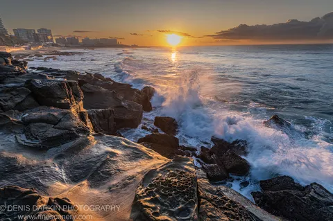 Honeycomb-weathered limestone at the Newcastle shoreline at dawn, a wave breaking at its base and the sun's reflection across the water.