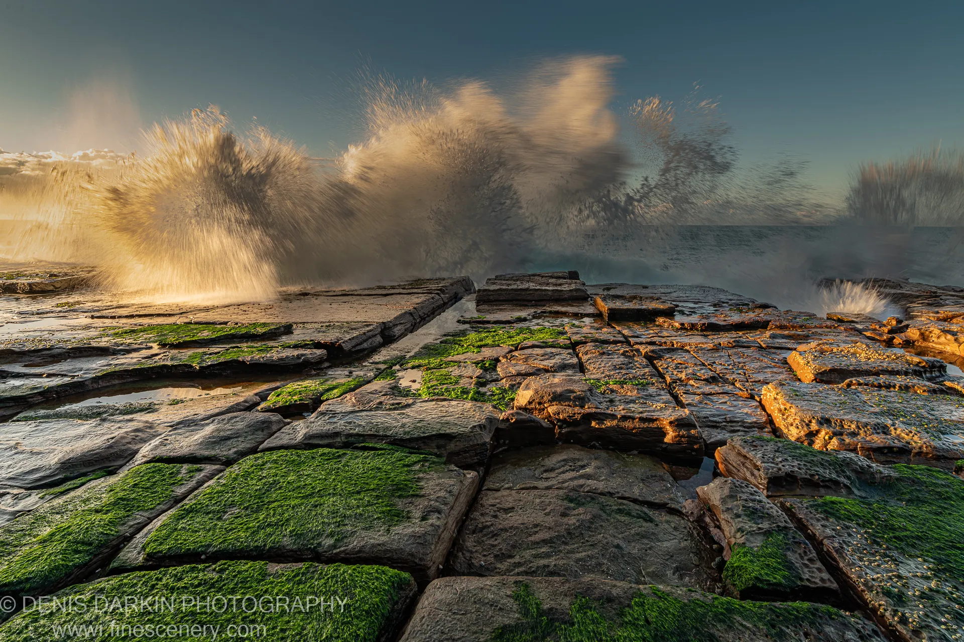 Sea spray erupts against dark stratified rock at Newcastle's coast, with green-moss-covered stone slabs in the foreground on a winter morning.
