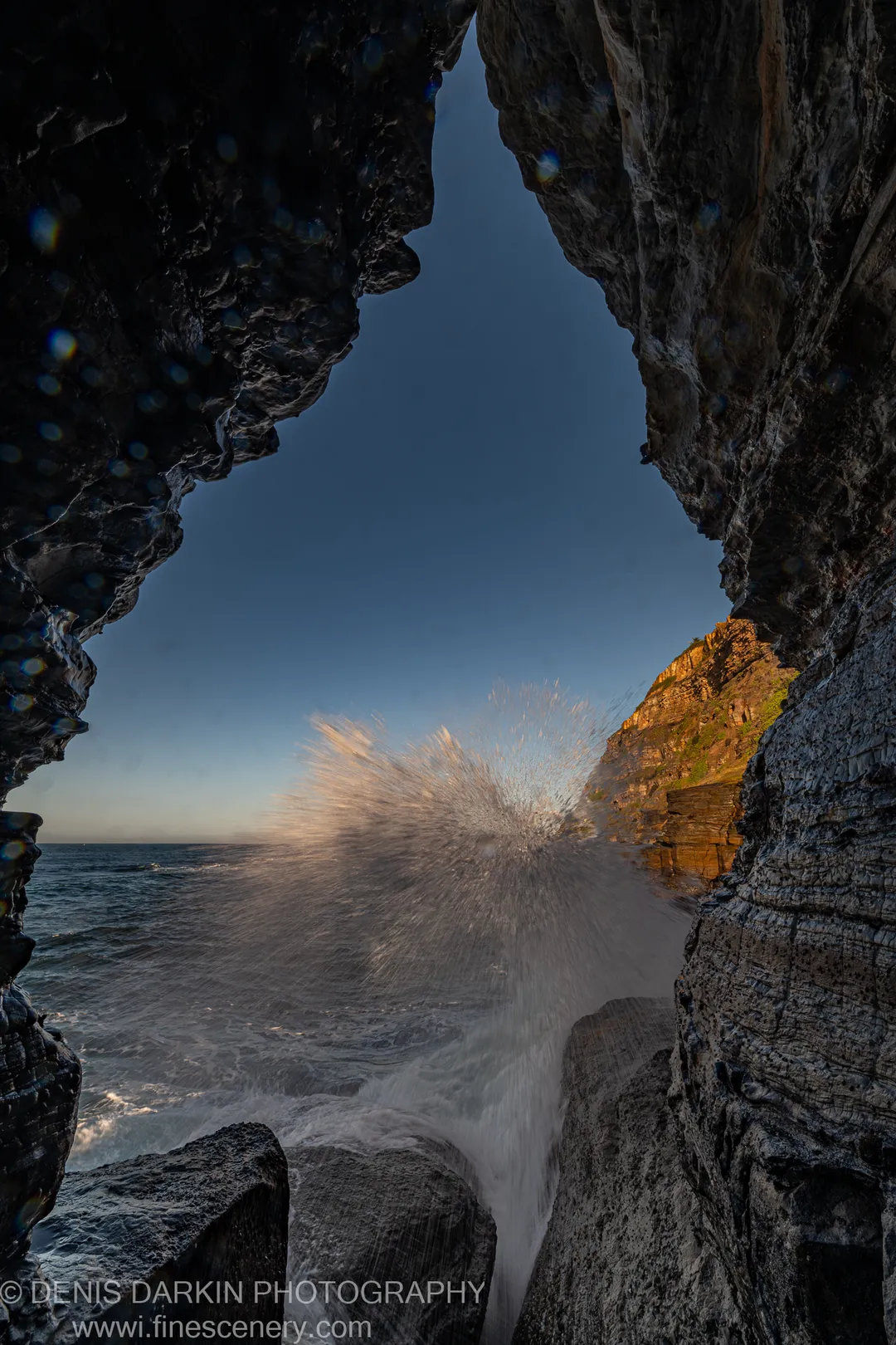 Wave erupting through a sea cave opening at Newcastle, with lens water droplets creating soft bokeh circles against the winter sky.