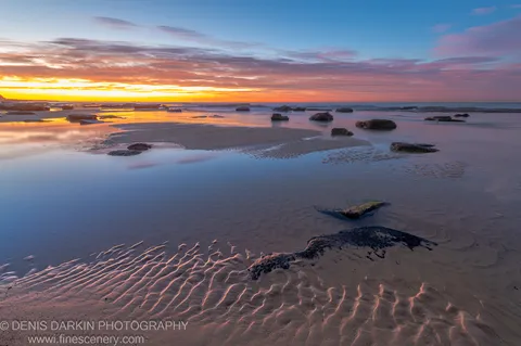 Scattered boulders and wet sand on the Newcastle coast at dawn, glassy long-exposure water between the rocks and a warm cloud-streaked horizon beyond.