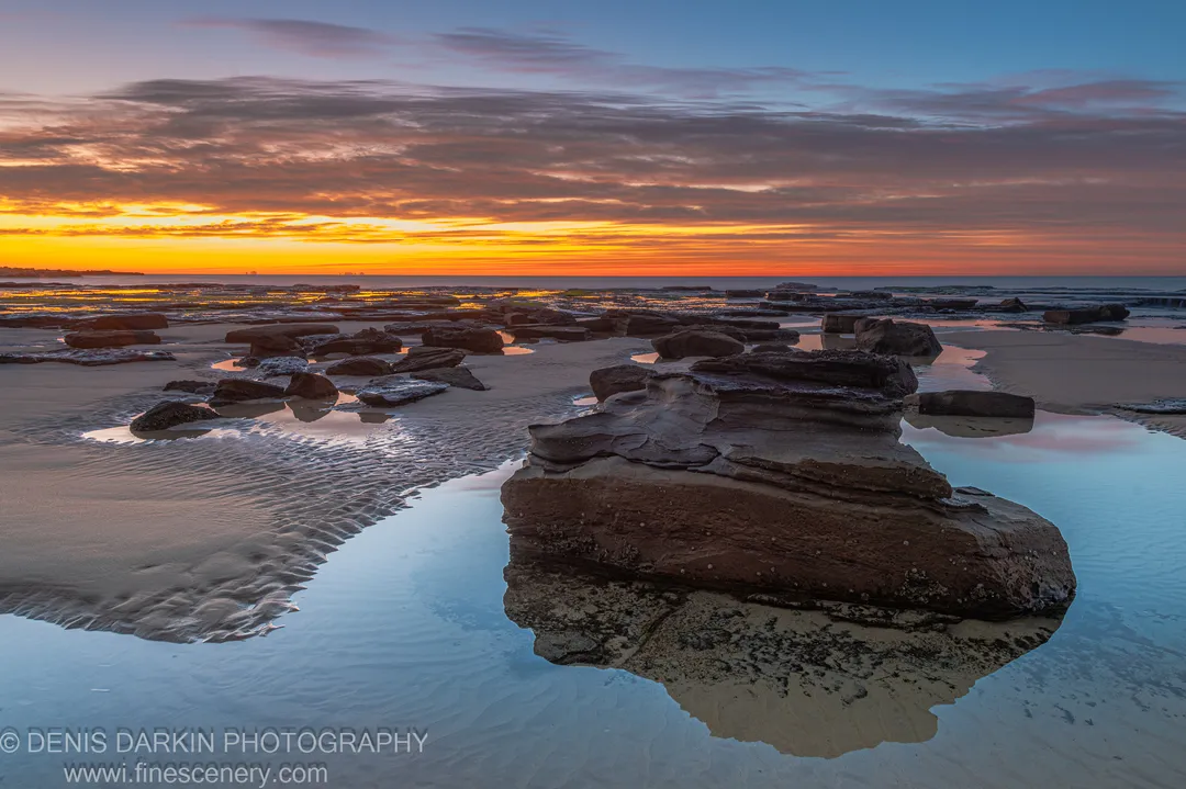 Layered sandstone in shallow tidal pools at dawn on the Newcastle coast, the still water reflecting warm horizon tones under a winter sky.
