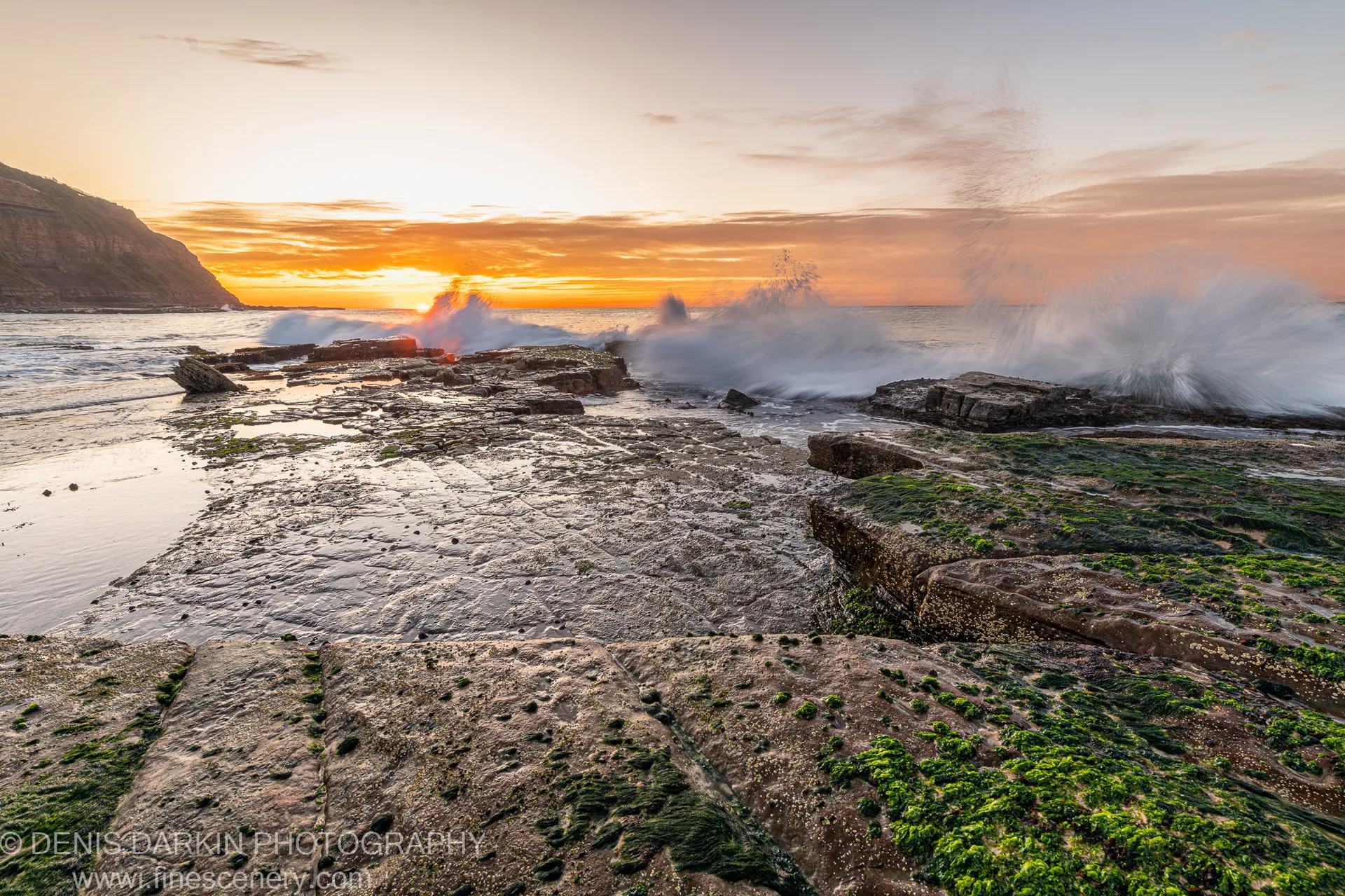 Algae-covered rock plates at Newcastle's coastline at dawn, crashing waves and sea spray visible at the horizon as sunlight breaks through winter cloud.