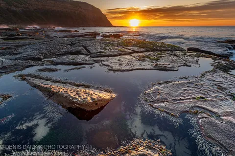 Layered sandstone cliff beside tidal pools reflecting the sunrise glow, with waves breaking on the distant shore at Newcastle, New South Wales.