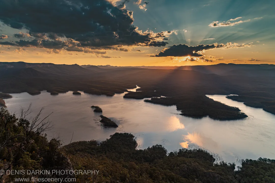 Crepuscular rays break through cloud above a winding lake at dusk, golden light reflected on calm water below forested hills, New South Wales.