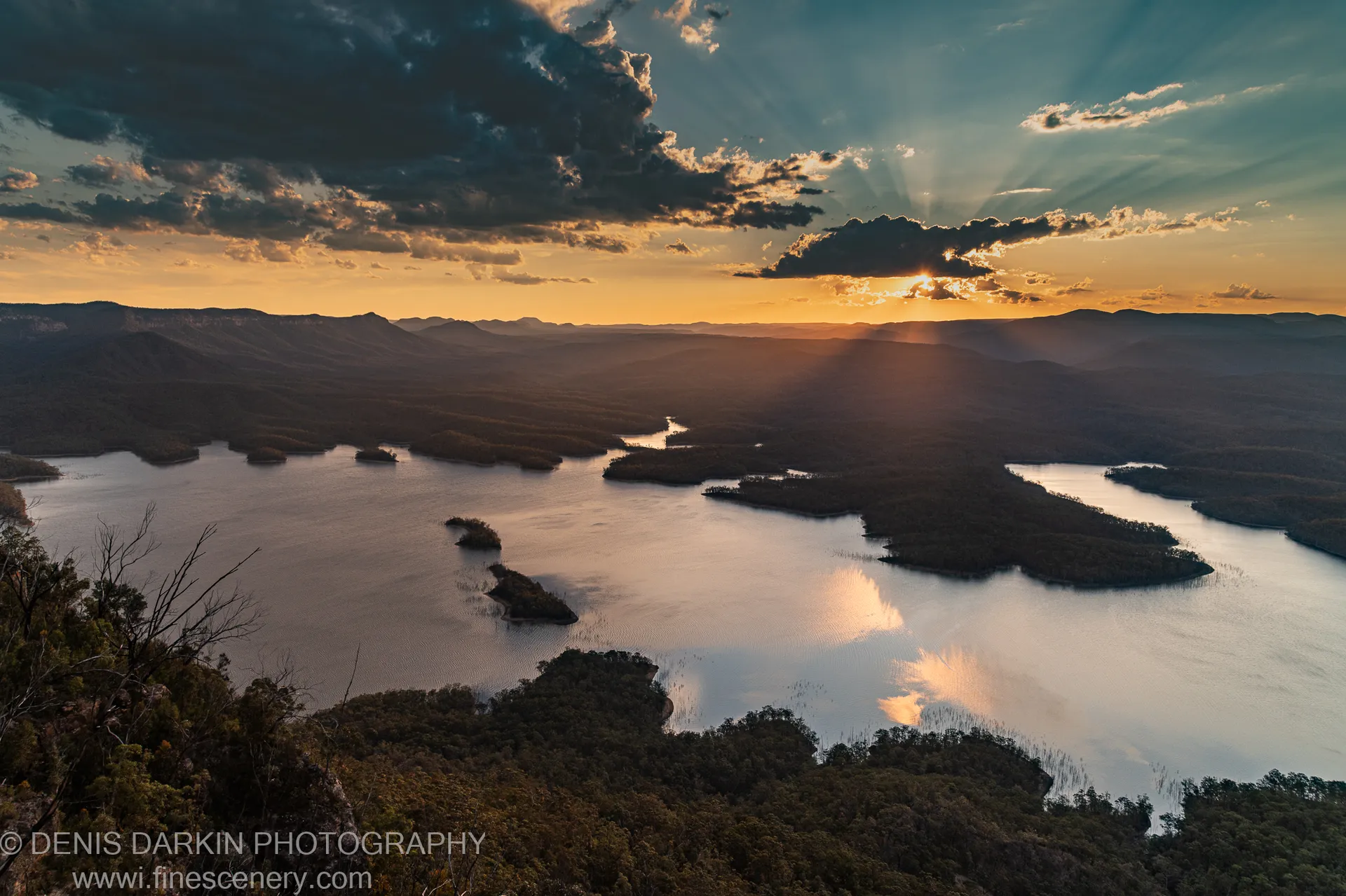 Crepuscular rays break through cloud above a winding lake at dusk, golden light reflected on calm water below forested hills, New South Wales.