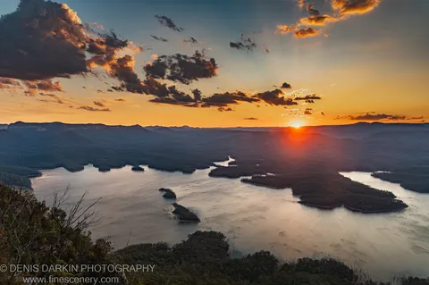 Evening sun breaks through heavy cloud above forested valleys and a winding river, casting golden reflections on calm water, New South Wales.