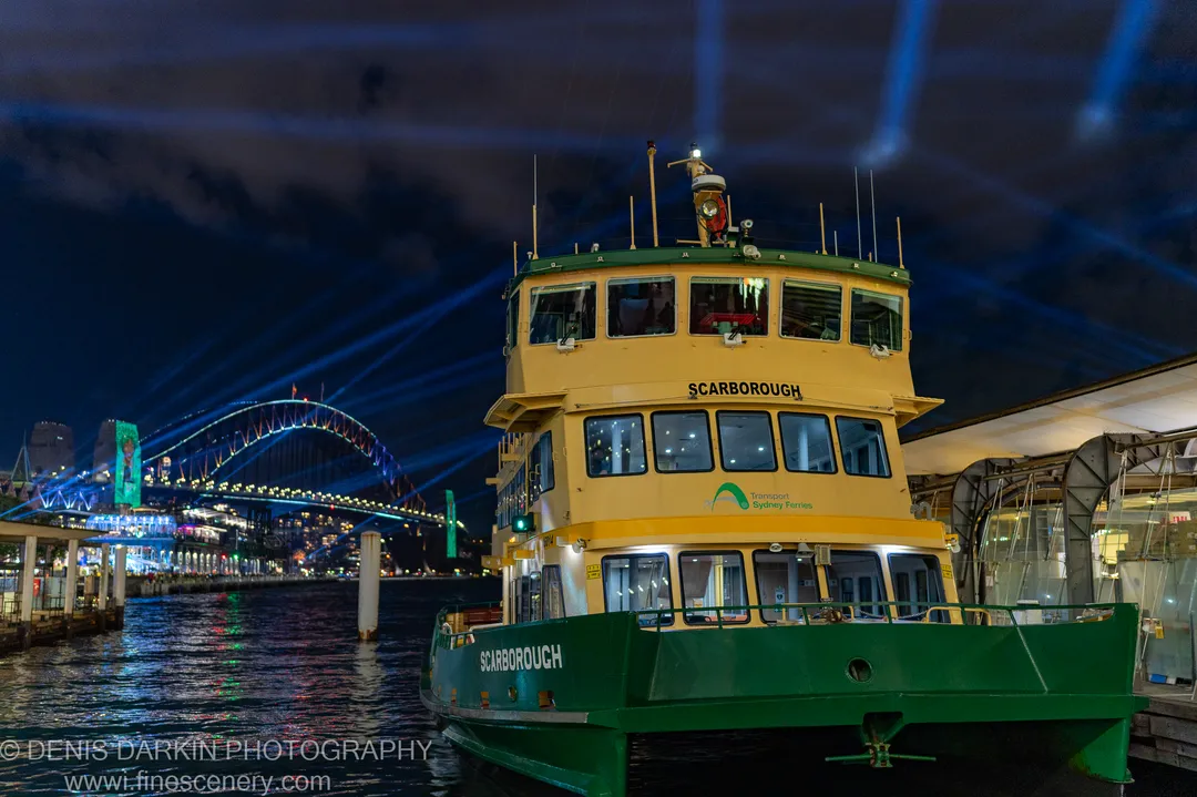 A yellow-and-green ferry sits docked at night in Sydney Harbour, the lit Harbour Bridge arch curving behind it against a dark sky.