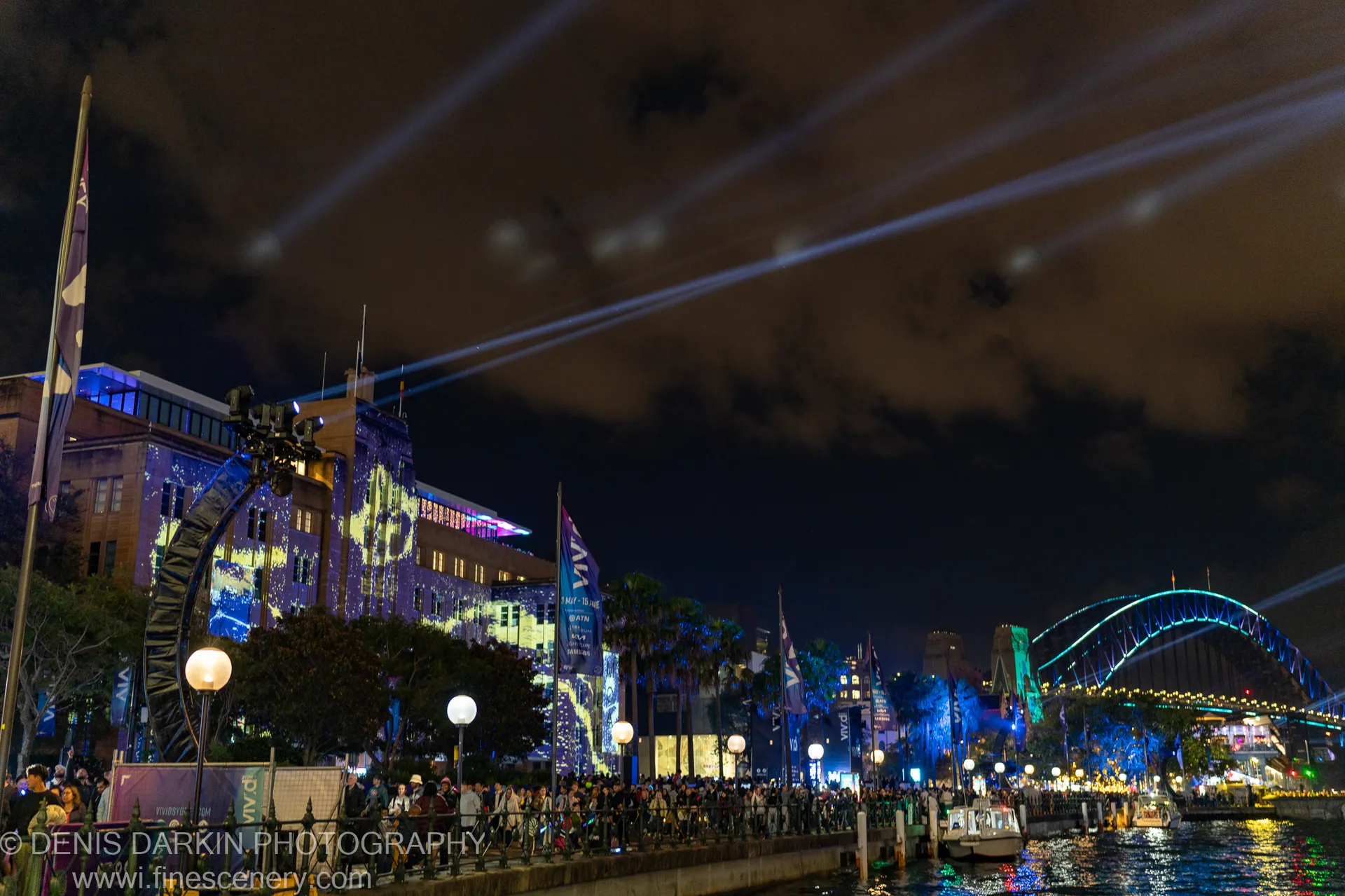 A crowd watches from Sydney's waterfront as diagonal searchlight beams and cyan bridge lighting fill the night sky.