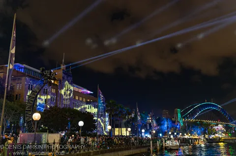 A crowd watches from Sydney's waterfront as diagonal searchlight beams and cyan bridge lighting fill the night sky.