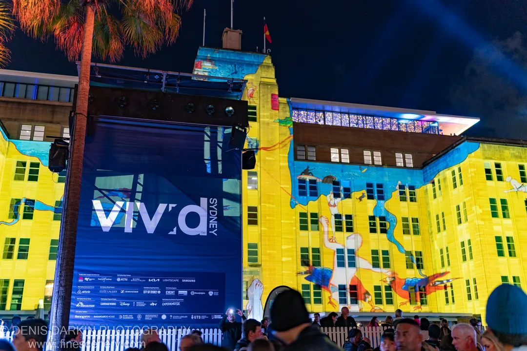 A historic sandstone building in Sydney serves as a night projection canvas, its warm stone surfaces carrying blue aquatic light patterns.