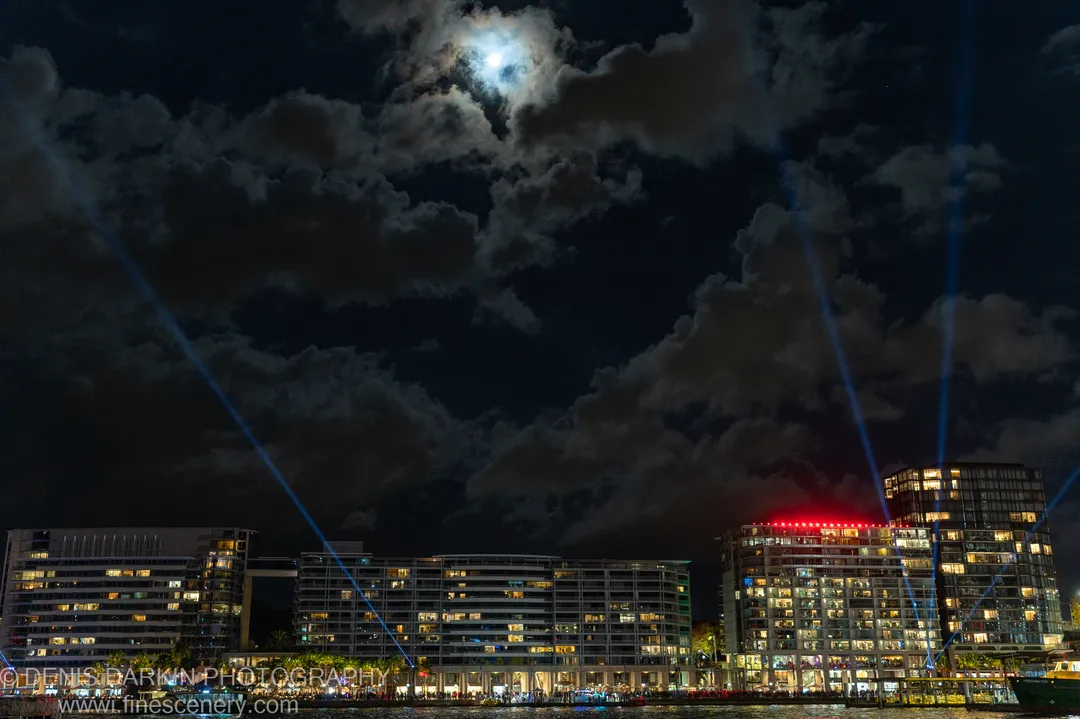 Blue laser beams rise diagonally through heavy cloud toward a bright moon over Sydney Harbour, with lit waterfront apartments lining the shore below.