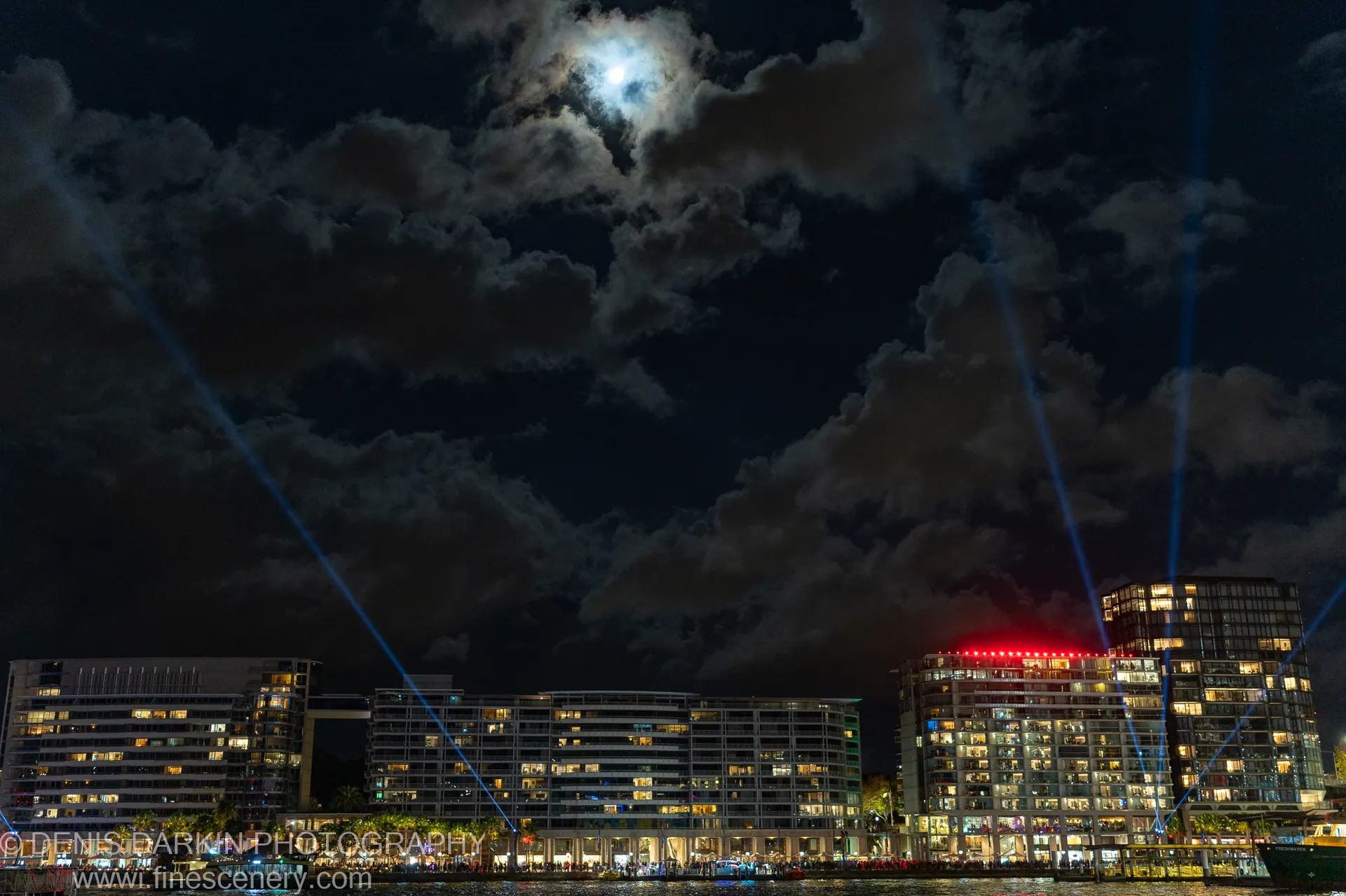 Blue laser beams rise diagonally through heavy cloud toward a bright moon over Sydney Harbour, with lit waterfront apartments lining the shore below.
