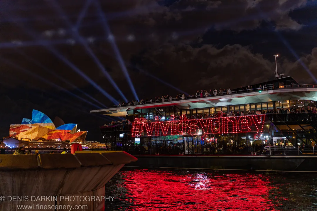 A barge carrying a red LED installation on Sydney Harbour, with blue spotlights overhead and the Opera House visible in warm light behind.