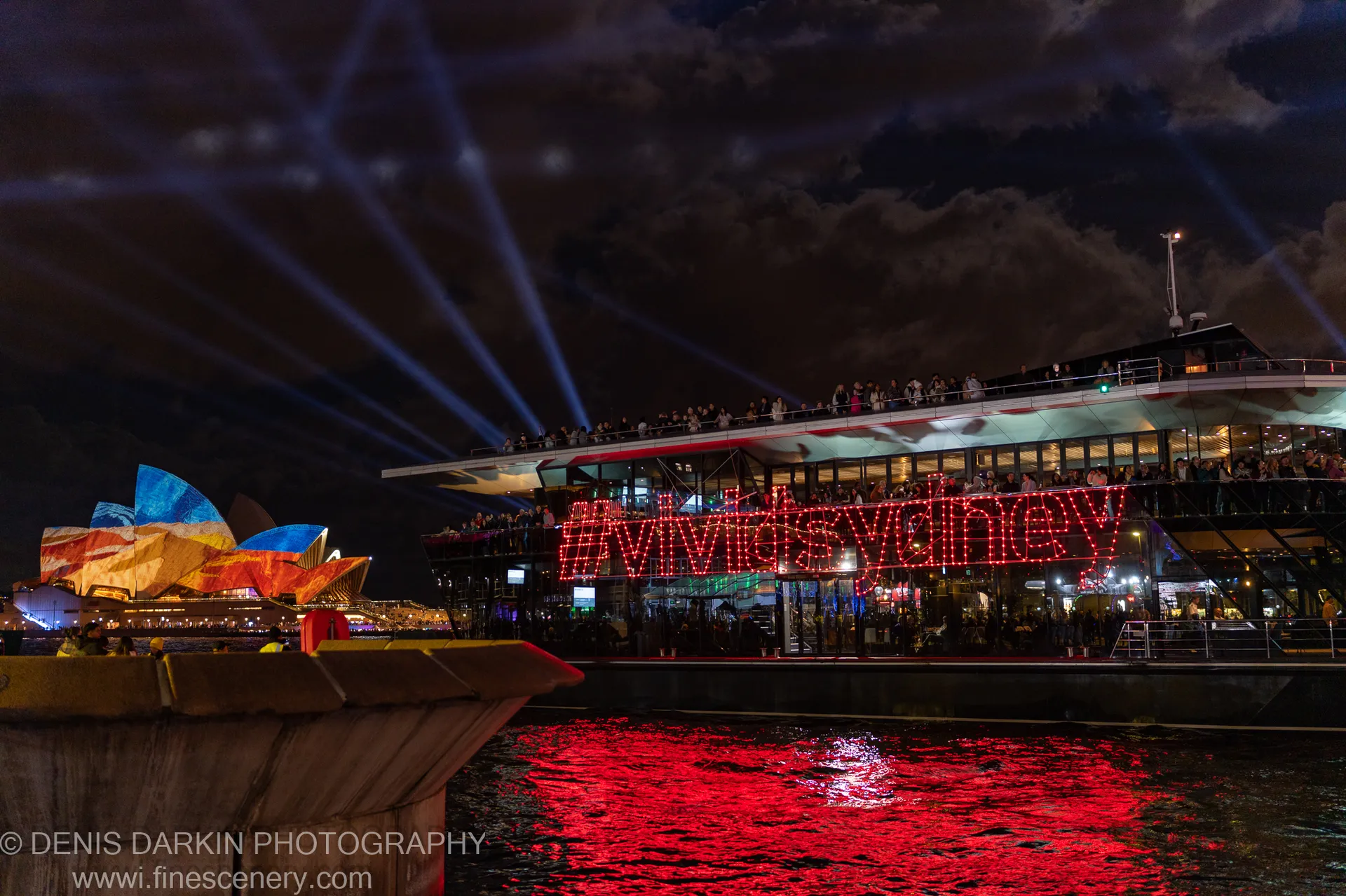 A barge carrying a red LED installation on Sydney Harbour, with blue spotlights overhead and the Opera House visible in warm light behind.