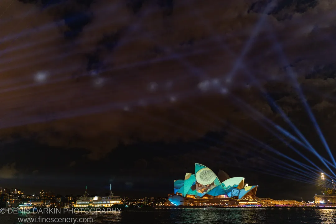 The Sydney Opera House wears a teal projection at night, blue spotlights rising into dark cloud while a ferry passes across the harbor.