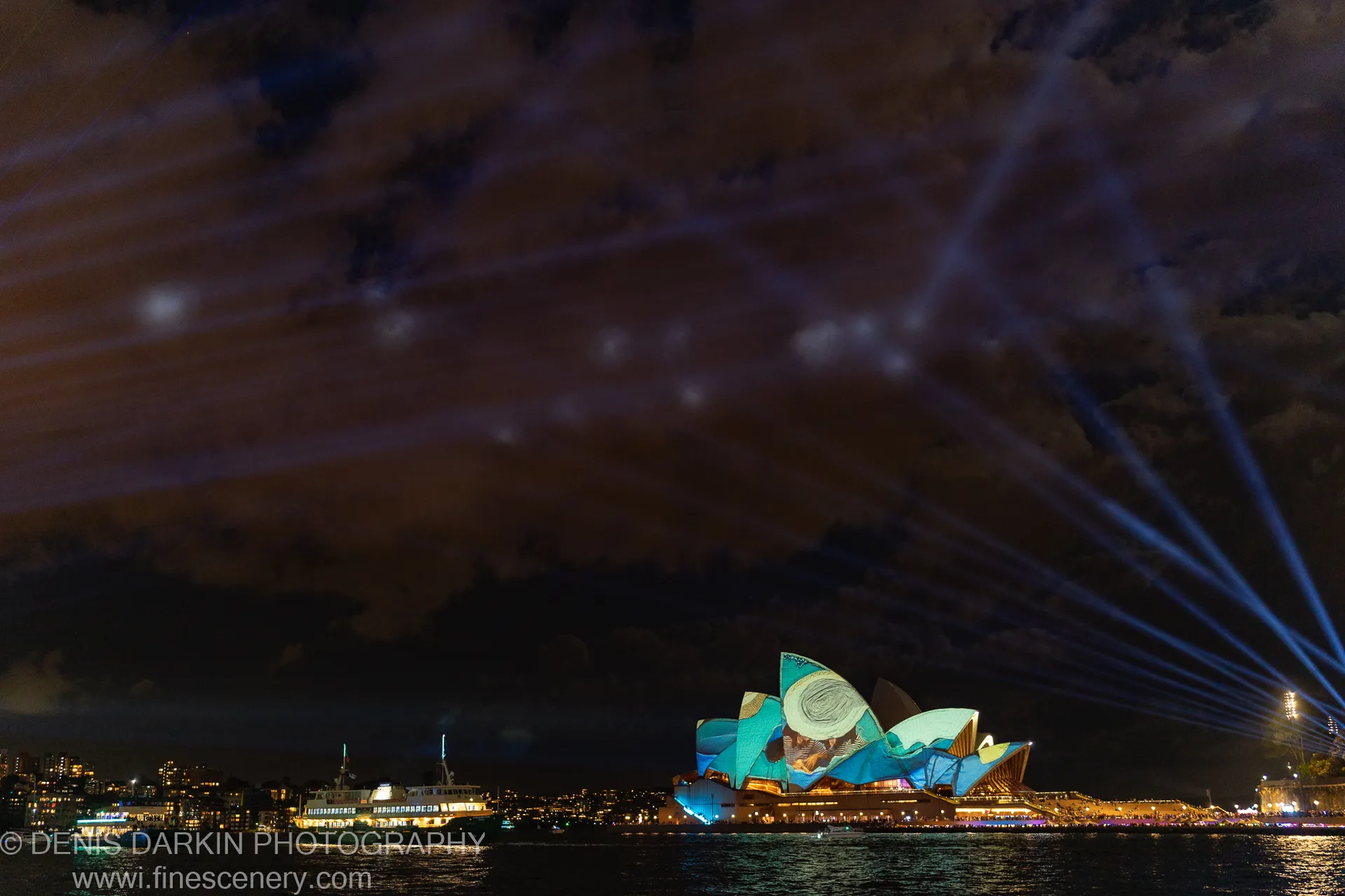 The Sydney Opera House wears a teal projection at night, blue spotlights rising into dark cloud while a ferry passes across the harbor.