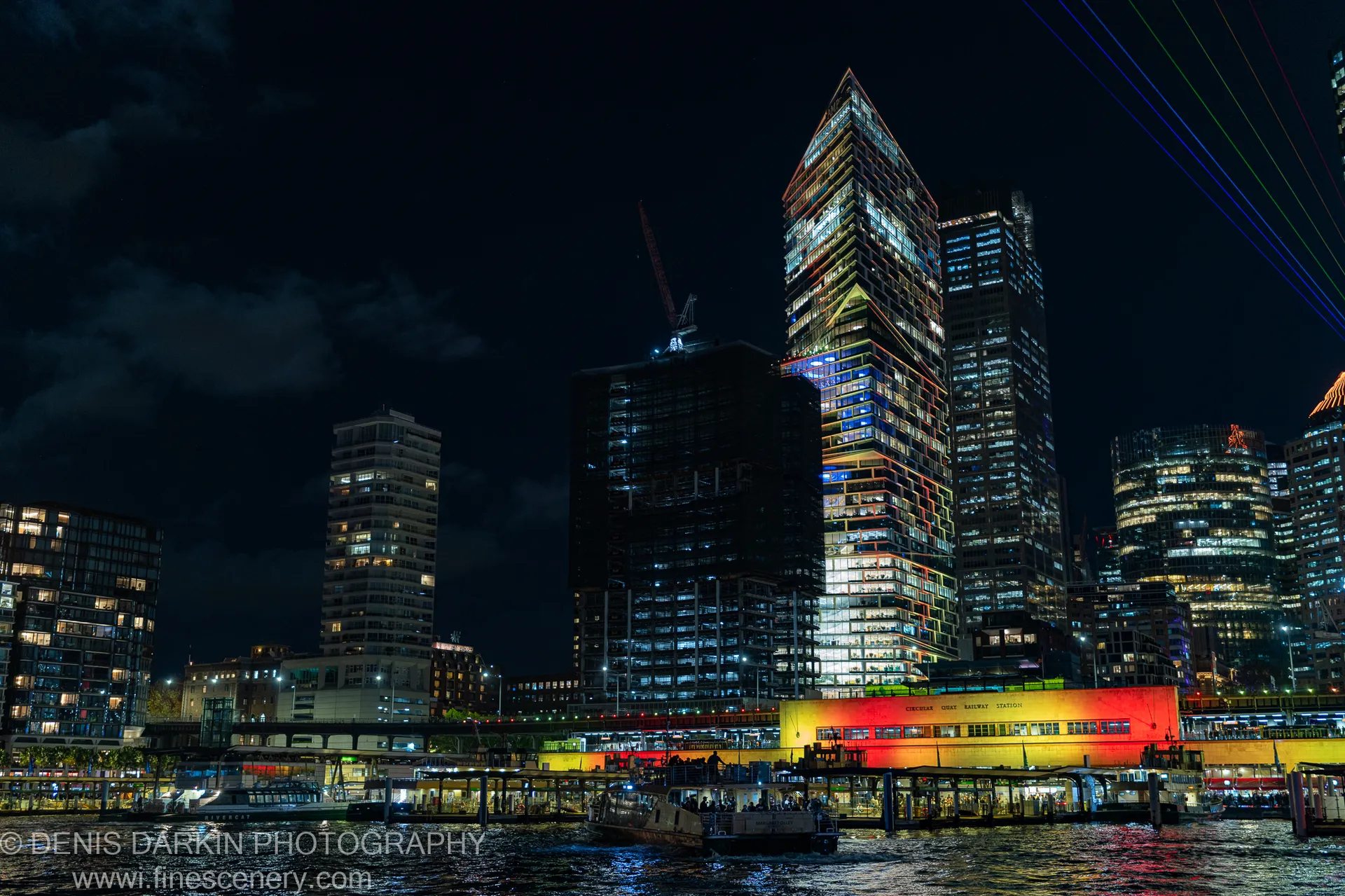 Sydney's waterfront towers at night, the central one carrying a neon gradient down its glass facade as a ferry crosses the dark harbor.