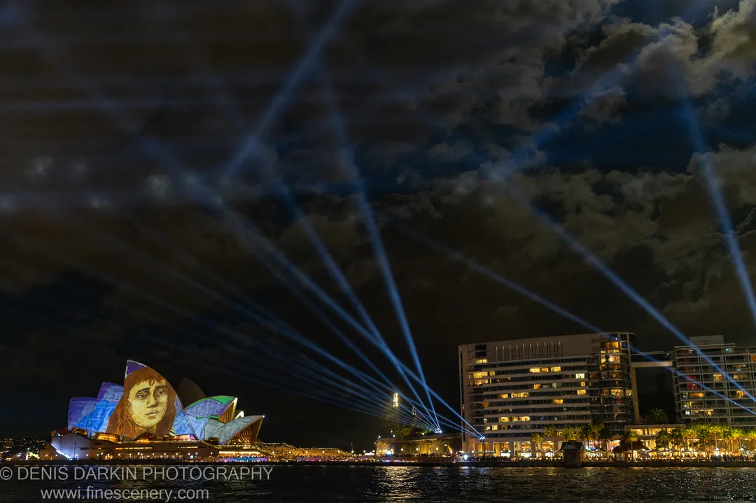 A colorful face projection covers the Opera House shell arches in Sydney as blue laser beams rise from the harbor edge.