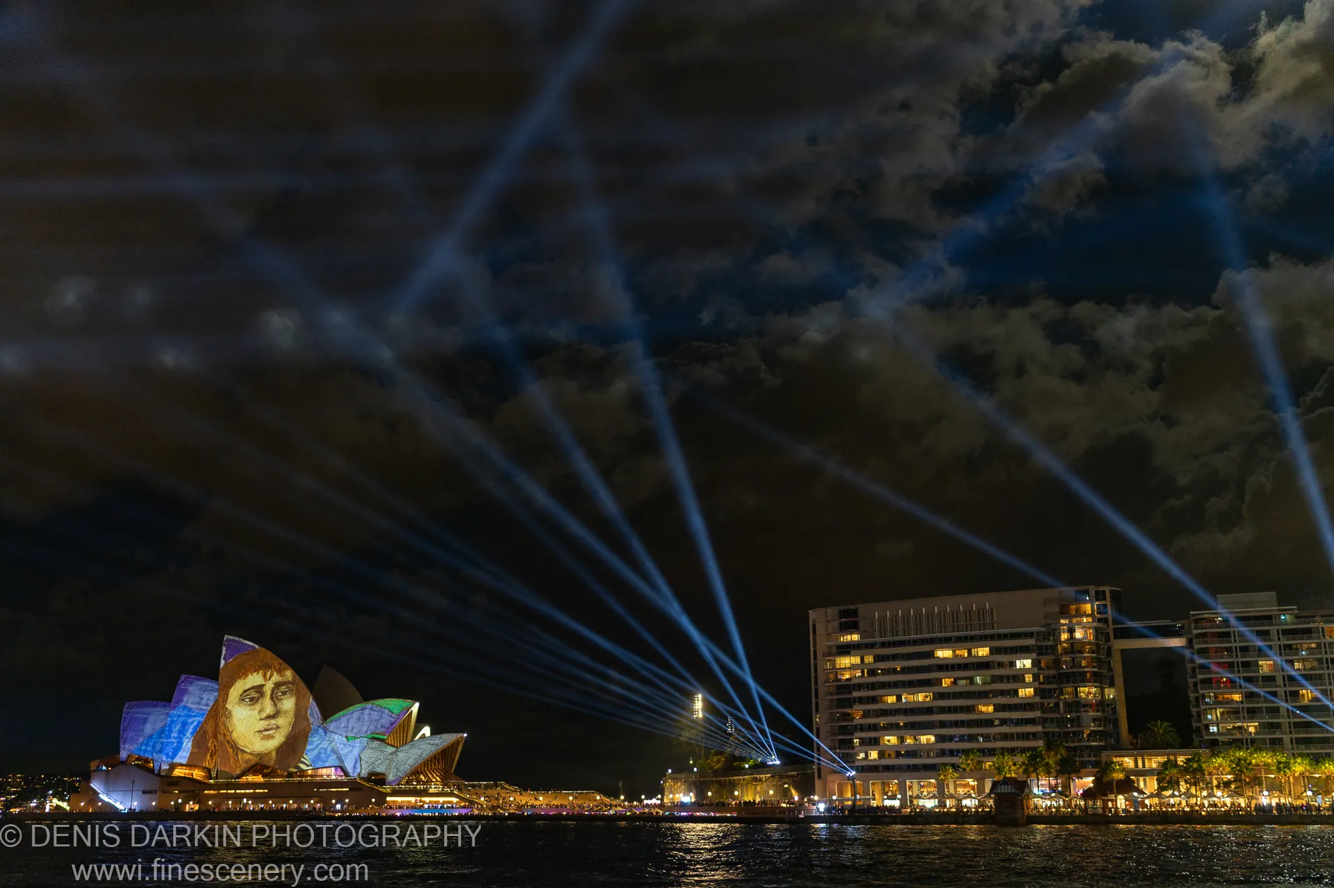 A colorful face projection covers the Opera House shell arches in Sydney as blue laser beams rise from the harbor edge.