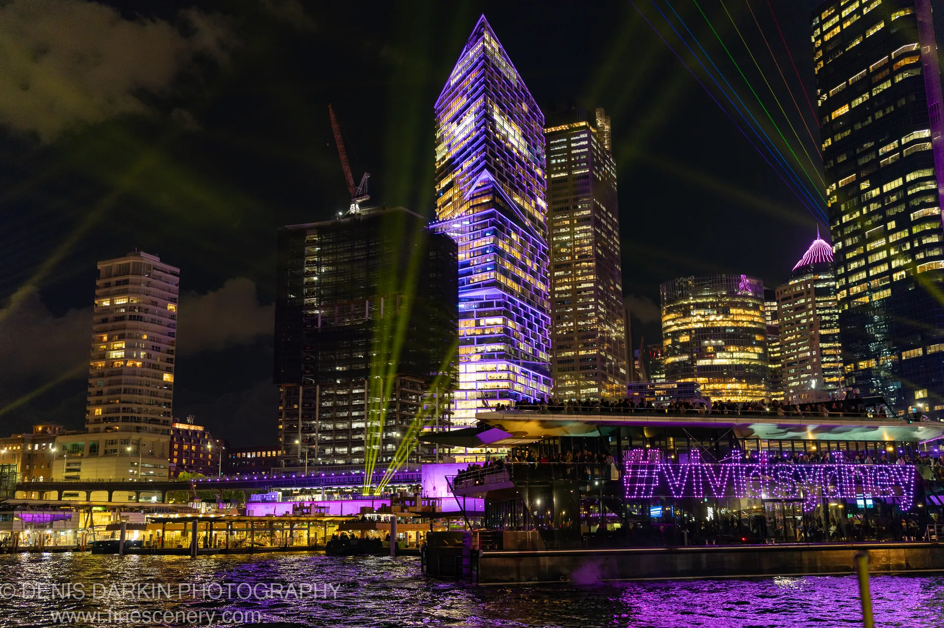 A skyscraper in purple and gold light on Sydney's waterfront at night, green laser beams crossing diagonally and reflecting on the water below.