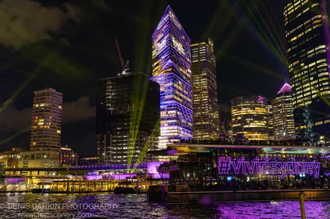 A skyscraper in purple and gold light on Sydney's waterfront at night, green laser beams crossing diagonally and reflecting on the water below.