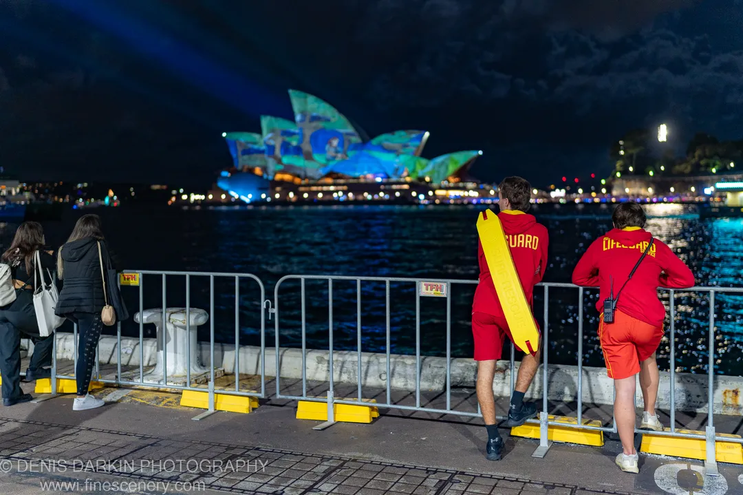 Two lifeguards in red uniforms lean on a railing in Sydney, watching light projections on the Opera House across the dark water.