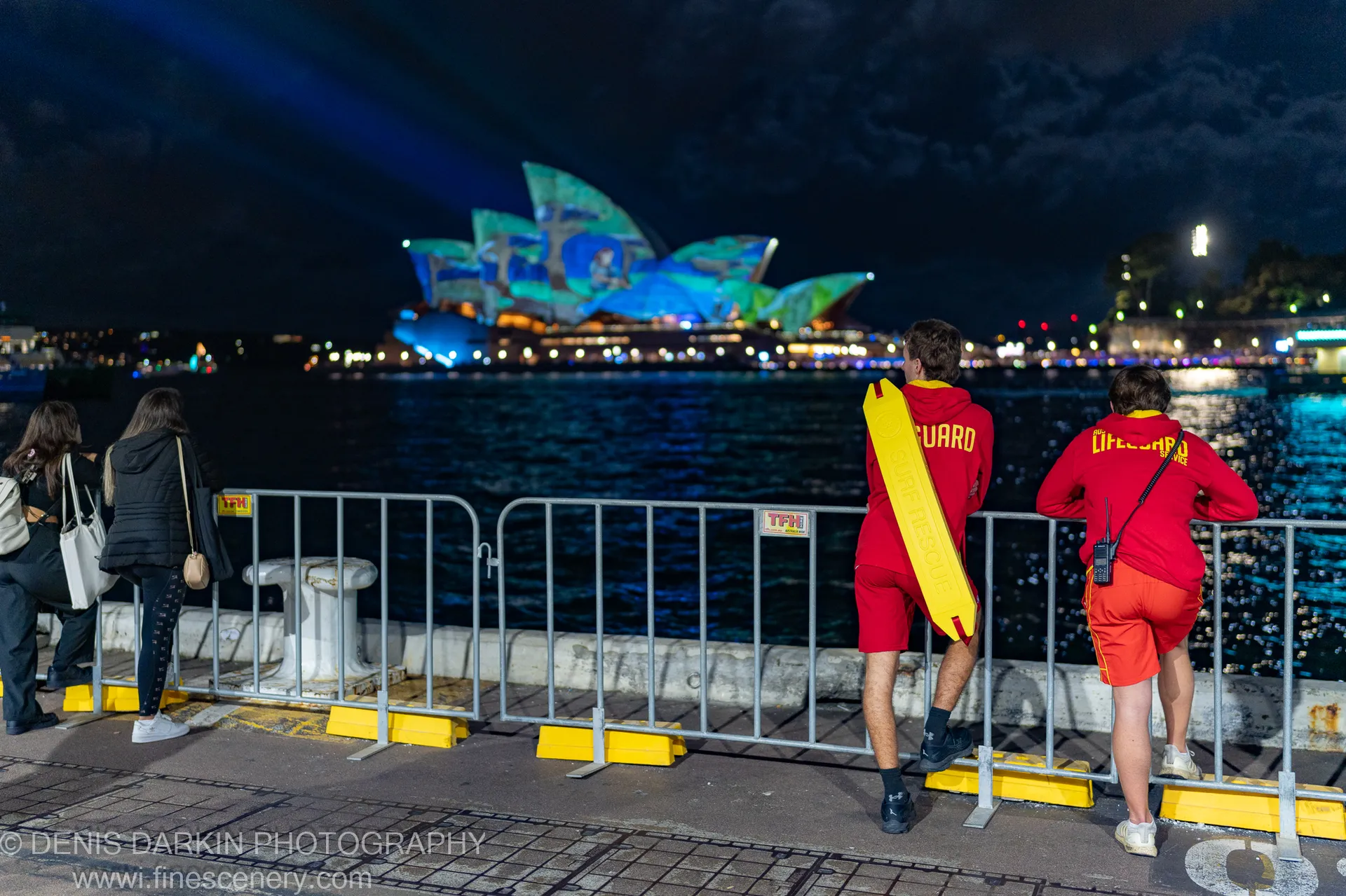 Two lifeguards in red uniforms lean on a railing in Sydney, watching light projections on the Opera House across the dark water.