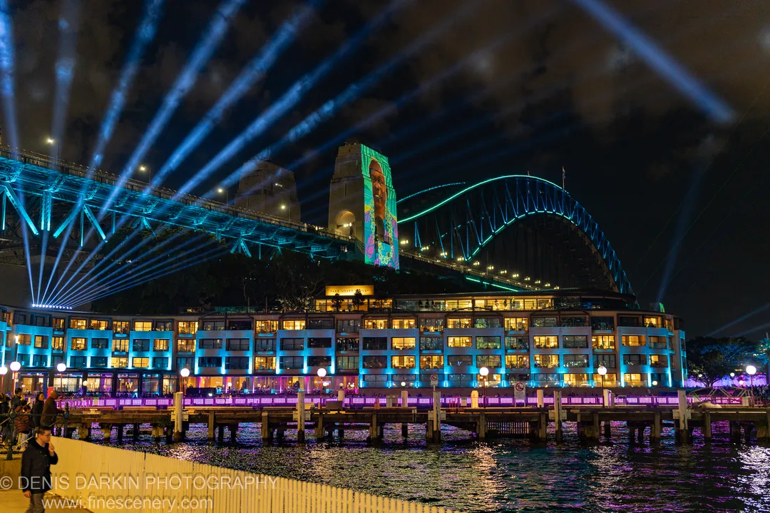 The Sydney Harbour Bridge lit with cyan lighting at night, with diagonal beams of light cutting through clouds above the waterfront.