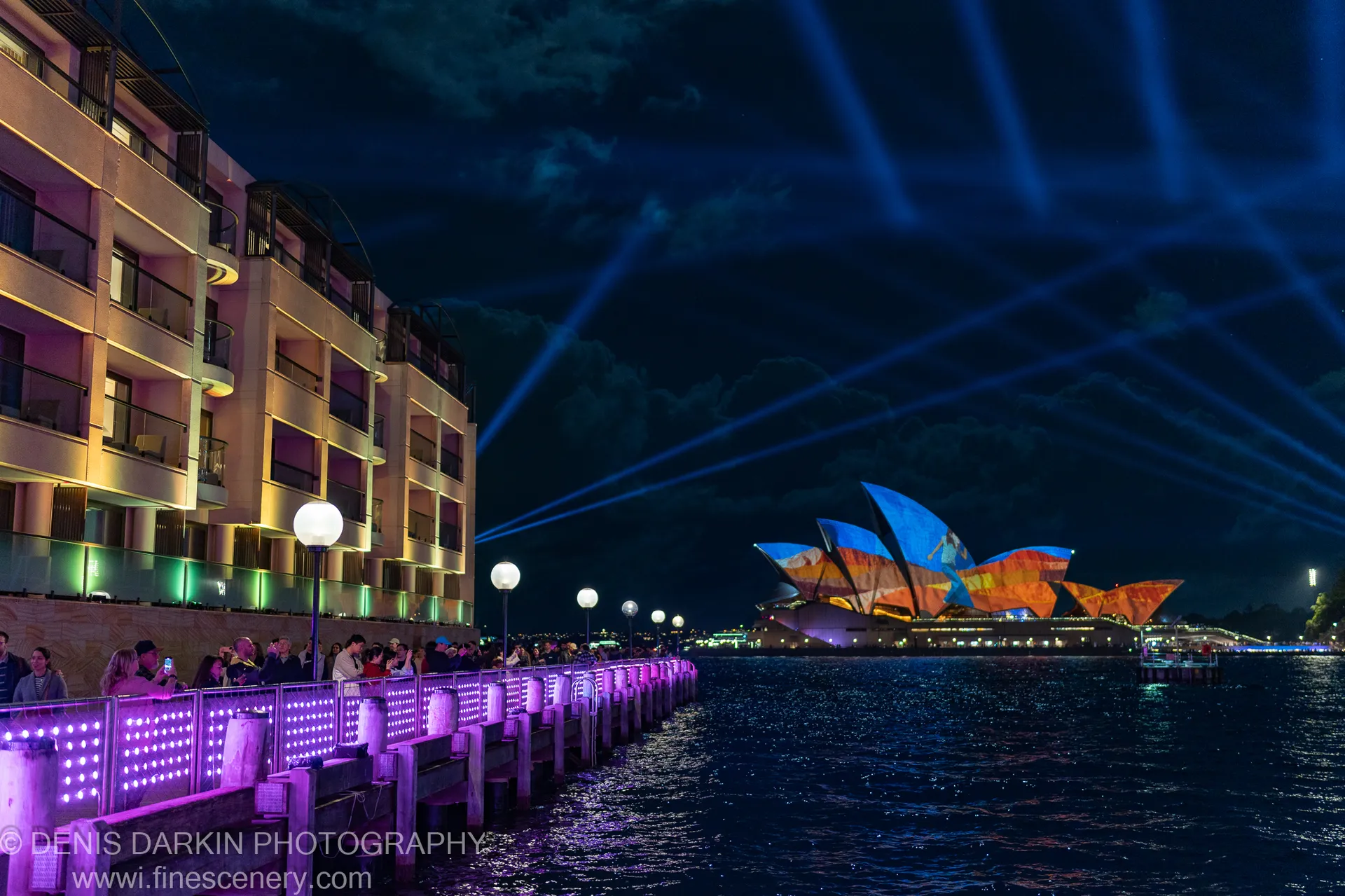 A wooden pier lined with purple LED lights curves past the illuminated Sydney Opera House under a night sky crossed by laser beams.