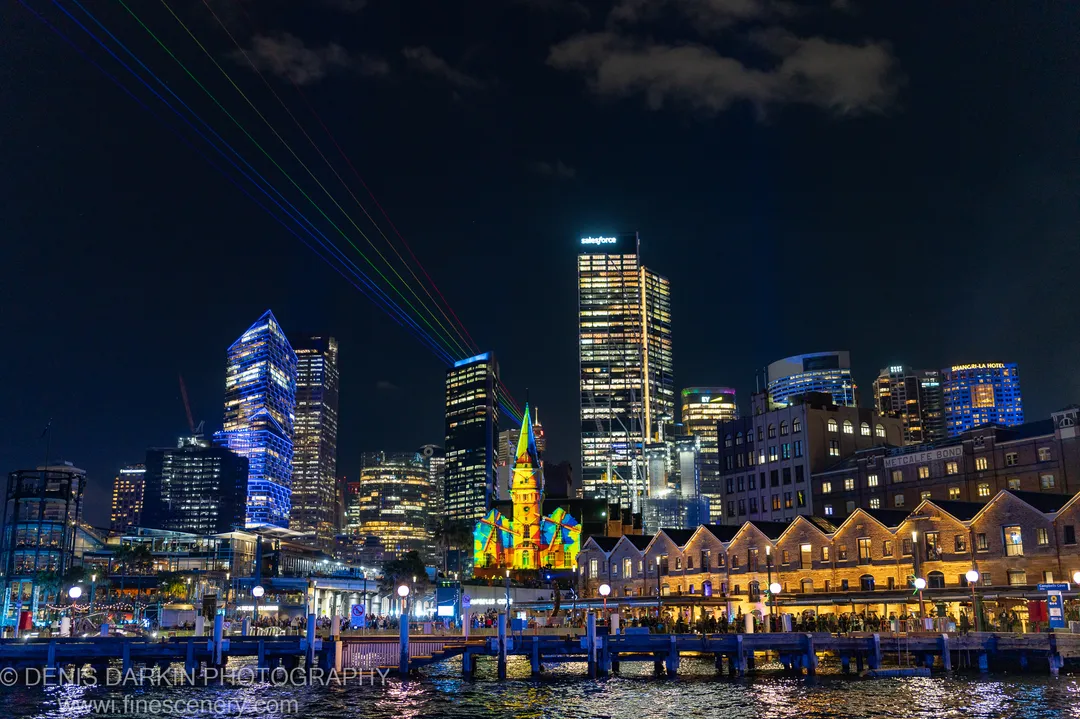 A historic sandstone church front covered in multicolored projection light, with laser beams crossing the night sky and lit towers behind it.