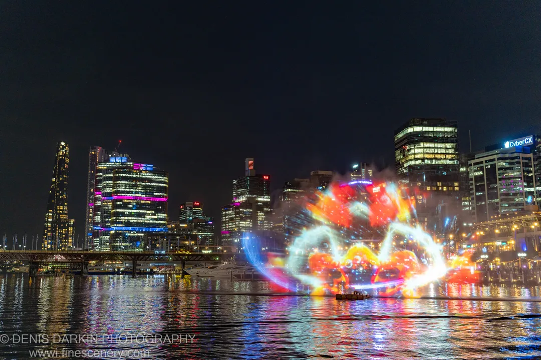 Night cityscape of Sydney Harbour with a bright orange and red water projection in the foreground, reflected against the lights of distant skyscrapers.