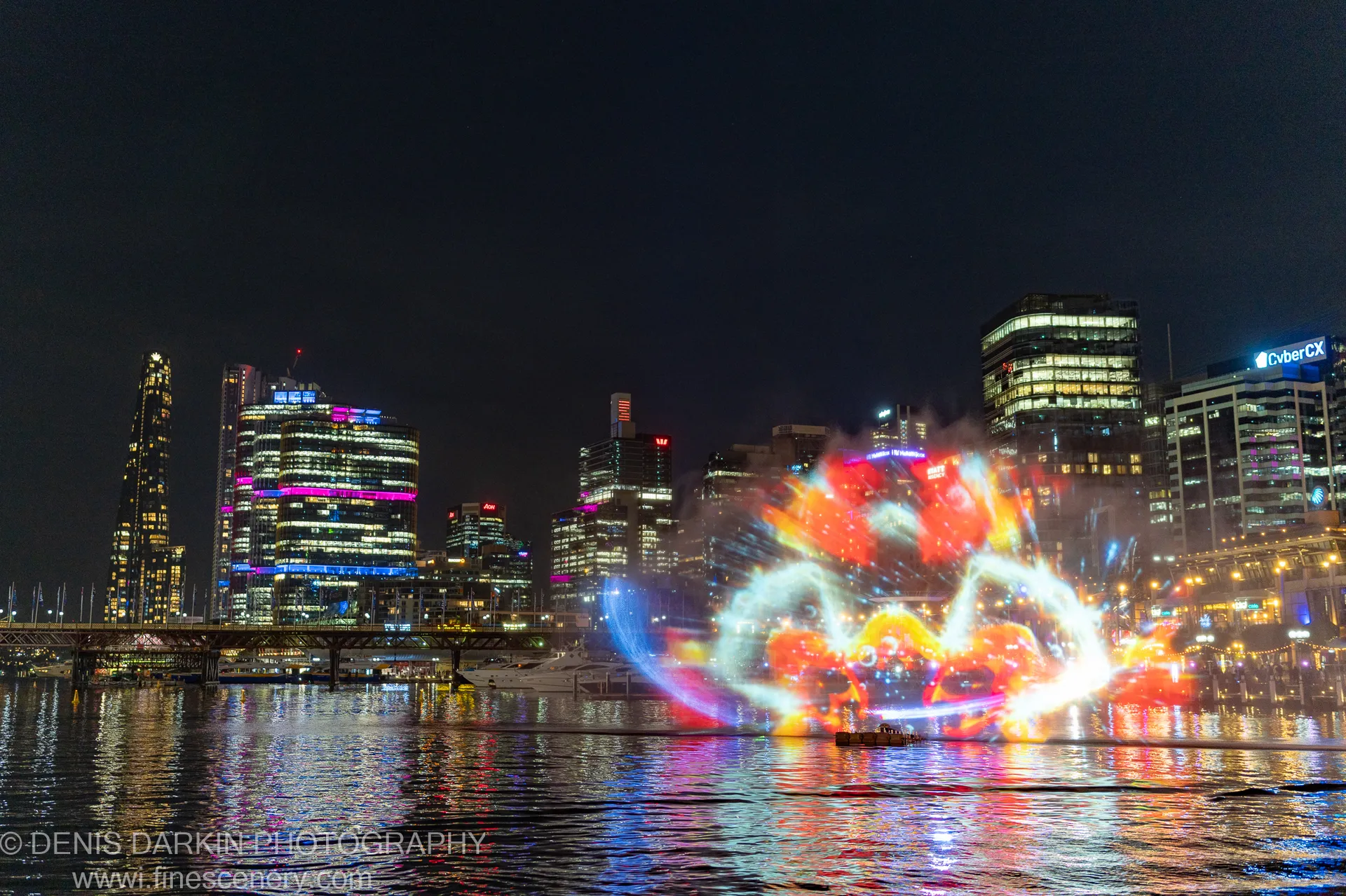 Night cityscape of Sydney Harbour with a bright orange and red water projection in the foreground, reflected against the lights of distant skyscrapers.