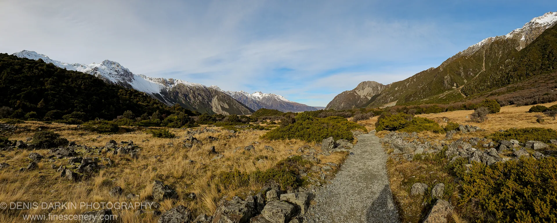 A gravel track winds through golden tussock and dark boulders toward snow-capped peaks under a clear blue sky, Central Otago.