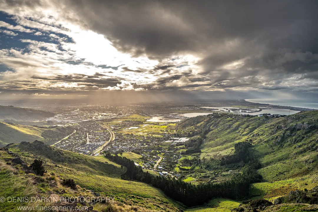 Distinct light beams break through dark clouds above a valley with urban sprawl and a coastal inlet, viewed from green slopes, Central Otago.